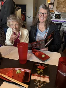 Two women sitting at a restaurant table with menus and empty sushi plates in front of them, smiling at the camera, enjoying a vibrant lifestyle and the sense of community that comes with sharing a meal together.