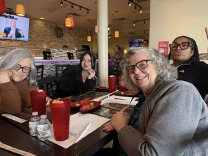 Four people sit at a restaurant table with menus, red drinks, and water bottles, smiling at the camera. Sushi dishes are on the table, reflecting a vibrant lifestyle and sense of community. A TV is on in the background.