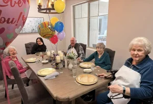 Five elderly people and one younger person sit around a dining table with balloons and birthday decorations, smiling and ready to celebrate in a warm display of community and joyful lifestyle.