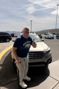 An older man with a cane stands in front of a parked Subaru, giving a thumbs up. He wears a "Nevada" t-shirt, sweatpants, and sneakers, embracing community and lifestyle in a sunny parking lot.