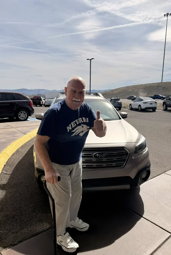 An older man with a cane stands in front of a parked Subaru, giving a thumbs up. He wears a "Nevada" t-shirt, sweatpants, and sneakers, embracing community and lifestyle in a sunny parking lot.