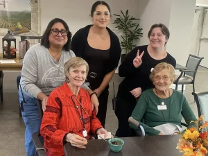 Five women, two seated and three standing behind them, pose and smile in a dining area with a table, flowers, and chairs visible, capturing the warmth of community and lifestyle.