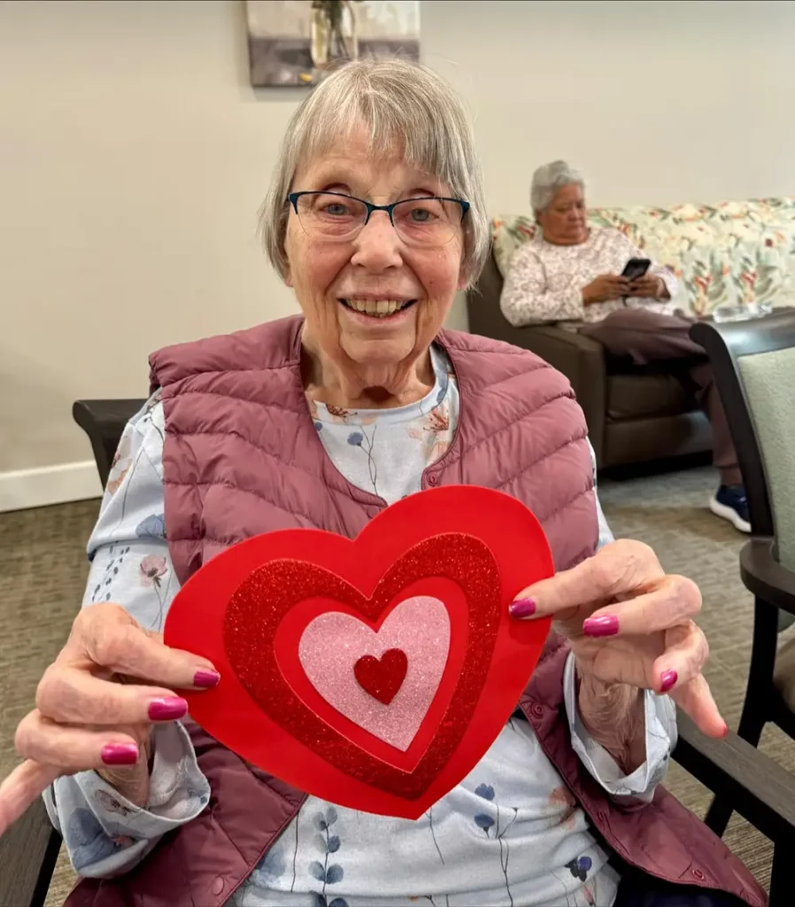 An elderly woman smiles while holding a large, handmade red heart decoration, reflecting a warm sense of community. Another woman sits in the background on a couch using a phone, embracing a relaxed lifestyle.