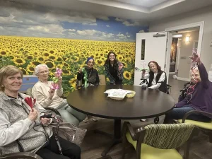 Six women sit around a round table in a room with a sunflower mural, holding up paper heart crafts and smiling at the camera, celebrating creativity and community.