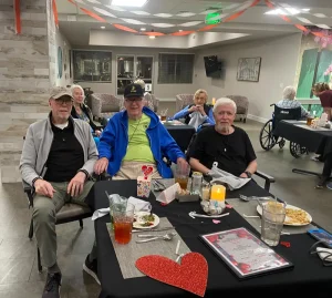 Three elderly men sit at a decorated table in a warm community dining area, enjoying food, drinks, and Valentine's Day decorations—a snapshot of shared lifestyle moments. Other people are seated in the background.