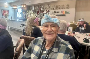 An elderly person wearing under-eye patches and a blue headband smiles, enjoying a vibrant community lifestyle while sitting in a communal dining area with others in the background.