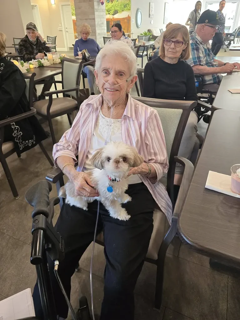 An elderly woman with white hair sits in a chair, smiling, with a small white dog on her lap. She enjoys the warm sense of community as others gather around her at tables in this friendly, shared setting.