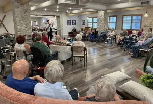 A group of elderly people sit in a large circle in a community room, engaged in an enriching lifestyle activity as they listen attentively to a speaker at the front of the room.