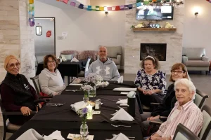 Six older adults sit around a dining table with a black tablecloth and folded napkins, enjoying community and lifestyle in a decorated room with flowers and a television on the wall.
