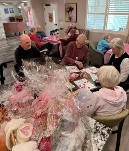 A group of seniors sit at tables playing bingo in a decorated room, enjoying a vibrant community lifestyle with pink gift baskets and balloons in the foreground.