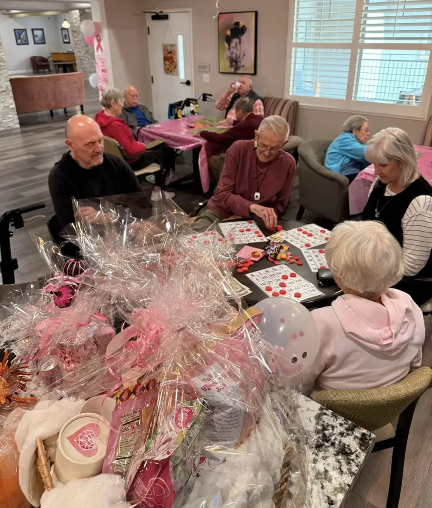 A group of seniors sit at tables playing bingo in a decorated room, enjoying a vibrant community lifestyle with pink gift baskets and balloons in the foreground.
