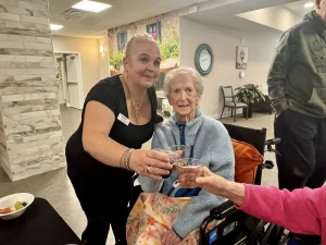 A woman and an elderly woman in a wheelchair clink plastic cups together in a joyful community celebration at an indoor event, highlighting the connections that enrich our lifestyle. Another person’s arm is extended to join the toast.
