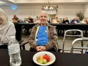 An elderly person sits at a table with a bowl of fruit and a water bottle in front of them, enjoying a healthy lifestyle amidst the warm atmosphere of a community room where others are seated in the background.