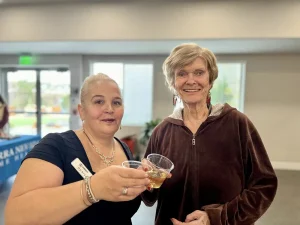 Two women standing indoors smile at the camera while holding plastic cups with drinks, toasting together and celebrating a moment of community in a well-lit room.
