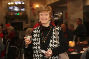 An older woman wearing a black and white argyle sweater holds a drink and a napkin at a festive indoor gathering, enjoying the lively community lifestyle with others in the background.