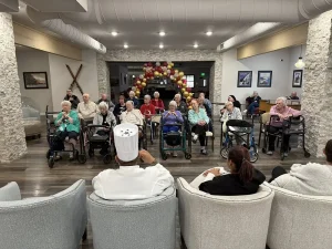 A group of elderly people sit in chairs and wheelchairs in a common room, enjoying a vibrant community lifestyle as they watch a chef and staff at the front. Red and gold balloons decorate the background.