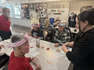 Elderly people sit at tables in a vibrant community space, wearing headbands and face masks, while staff assist them with a skincare activity that promotes healthy lifestyle habits.