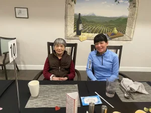 Two women sit side by side at a dining table with placemats and drinks, enjoying a relaxed lifestyle; a vineyard mural creates a warm sense of community on the wall behind them.