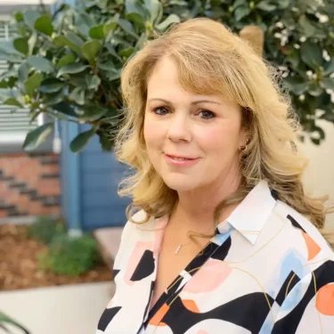 Sabrina Boyle, with blonde hair, stands outdoors in front of a leafy green tree, wearing a white patterned blouse and smiling slightly at the camera.