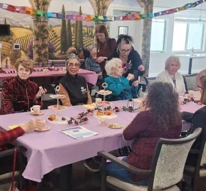 A group of elderly women sit around a table with tea and pastries, decorated with pink tablecloths and flowers; caregivers stand nearby in a brightly lit room.