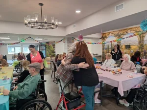 A group of elderly people and staff gather in a decorated room for a celebration; two women embrace near a table with snacks.
