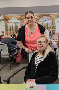 A woman stands behind another woman who is seated at a table in a communal dining area with several people in the background.