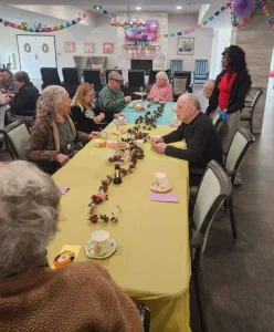 A group of elderly people sits around decorated tables having tea and conversation in a brightly decorated communal room.