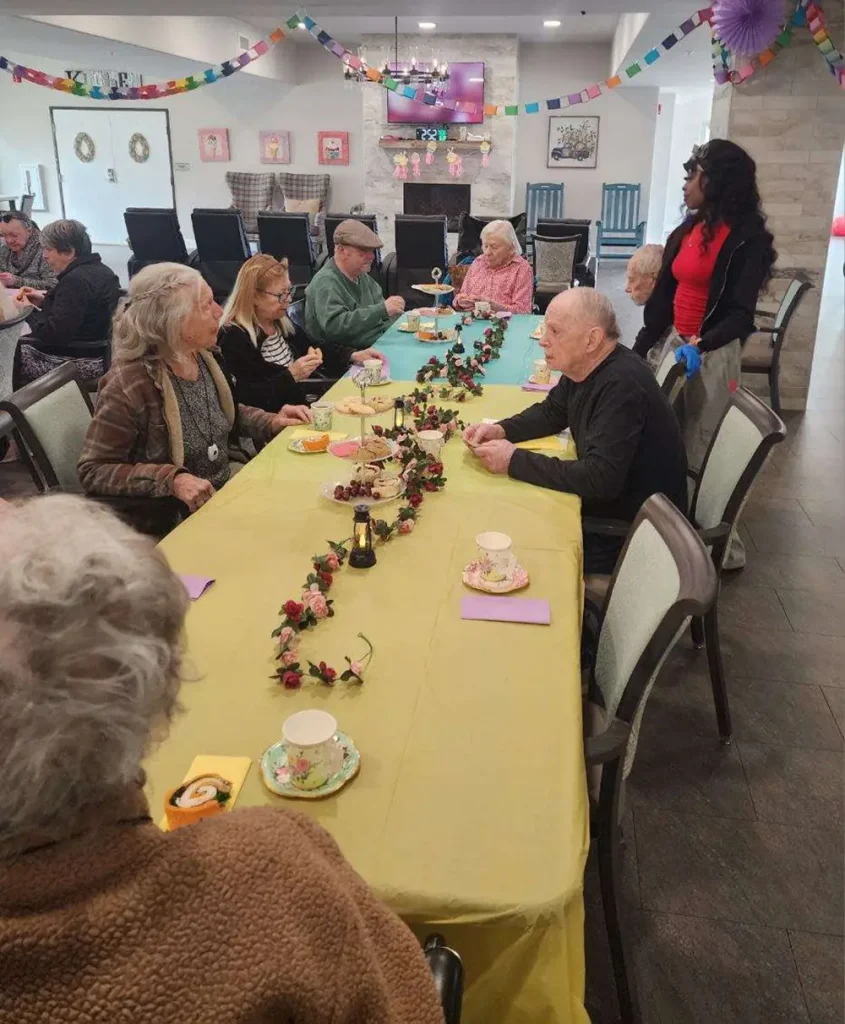A group of elderly people sits around decorated tables having tea and conversation in a brightly decorated communal room.