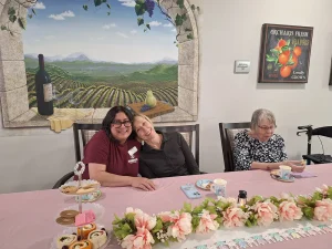Three women sit at a decorated table with pastries and drinks; one woman hugs another while the third sits nearby. A vineyard mural is on the wall behind them.