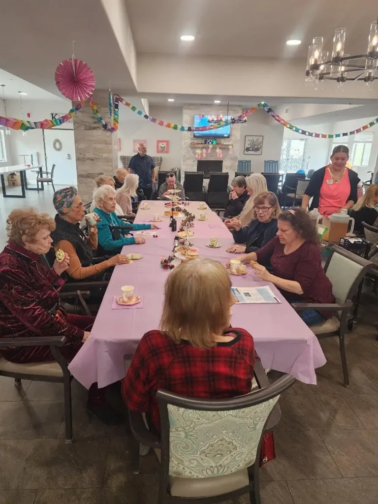 A group of elderly women sit around a long table with a pink tablecloth, having tea and snacks in a decorated room. Other people stand and chat in the background.