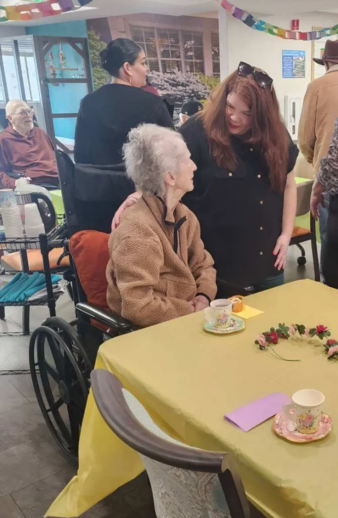 An elderly woman in a wheelchair sits at a decorated table, talking to a standing woman at a social gathering with other people in the background.