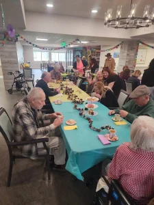A group of elderly people and caregivers sit around a decorated table, eating and socializing in a brightly lit room with festive decorations.