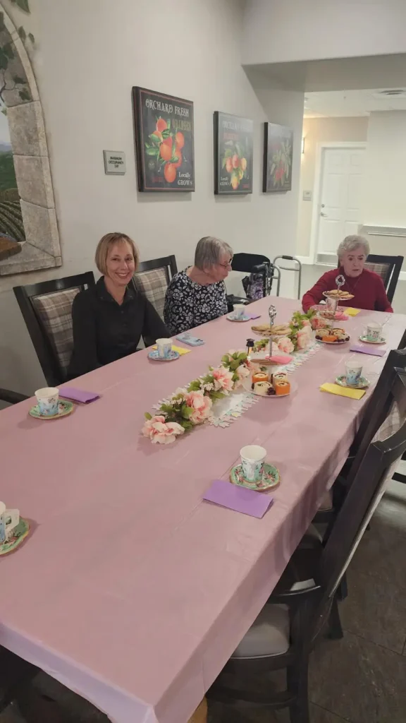 Three women are seated at a long table with a pink tablecloth, floral centerpiece, and teacups, set for a gathering in a bright indoor space.