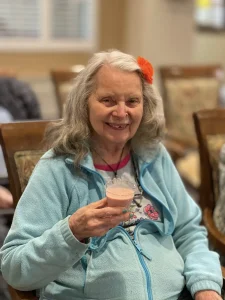 An older woman with long gray hair and a flower in her hair sits on a chair, smiling and holding a cup with a pink beverage. Wearing a light blue jacket, she radiates positivity, embodying health awareness on World Health Day.