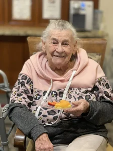 An elderly woman with gray hair, wearing a pink and leopard-print hoodie, sits in a chair holding a small plate of fruit and dessert, smiling at the camera—embracing health awareness on World Health Day.