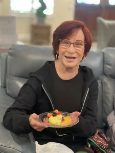 An older woman with short red hair and glasses sits on a gray couch, smiling and holding a dessert plate with fruit and cake—celebrating health awareness in her daily routine.