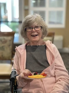 Older woman with gray hair and glasses, wearing a pink jacket, smiling and holding a plate with fruit slices while seated indoors—embracing health awareness in her daily life.