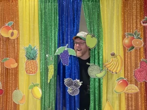 A man smiling and standing behind colorful, glittery curtains decorated with various paper fruit cutouts, celebrating World Health Day.