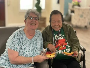 Two older women sitting indoors, smiling at the camera and holding plates with sliced fruit—celebrating health awareness on World Health Day.