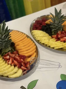 Two metal trays on a table display sliced cantaloupe, pineapple, strawberries, raspberries, and blackberries—an inviting spread perfect for World Health Day or boosting health awareness. A plastic tong lies nearby.