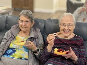 Two elderly women sit on a couch, smiling and holding plates of cut fruit. One woman holds a fork with a piece of fruit, celebrating health awareness and making mindful choices for global health.