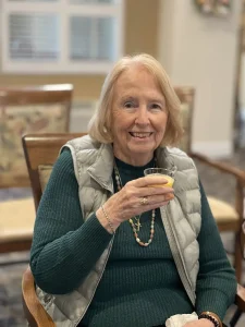 An older woman sits on a wooden chair indoors, smiling and holding a glass of orange juice. She wears a green sweater, gray vest, and beaded necklace, embodying health awareness in honor of World Health Day.