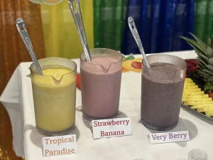 Three pitchers filled with different smoothies—Tropical Paradise, Strawberry Banana, and Very Berry—each with a serving spoon, are displayed on a table to celebrate World Health Day and promote nutritious choices.