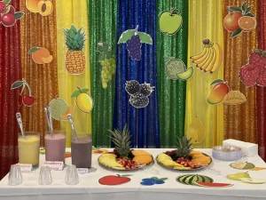 A colorful fruit-themed party table, perfect for World Health Day, features three smoothies, fruit platters, and vibrant fruit decorations set against rainbow sequin curtains in the background.