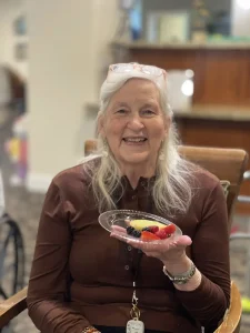 An older woman with long gray hair, wearing a brown shirt, smiles while holding a plate of assorted fruit indoors—celebrating World Health Day and promoting health awareness through nutritious choices.