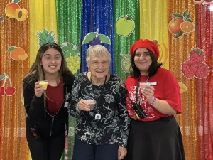 Three people stand in front of a colorful fruit-themed backdrop, smiling and holding drinks, as they celebrate World Health Day—with two younger women on either side of an elderly woman, promoting health awareness across generations.
